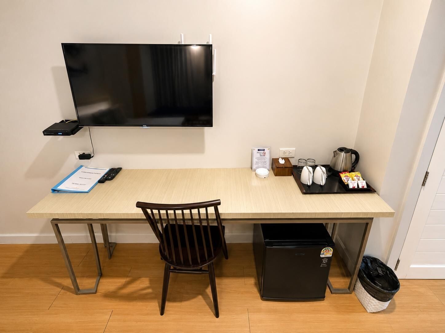 Una habitación pequeña y moderna con escritorio, silla, televisor, refrigerador miniatura y un cesto de basura.
