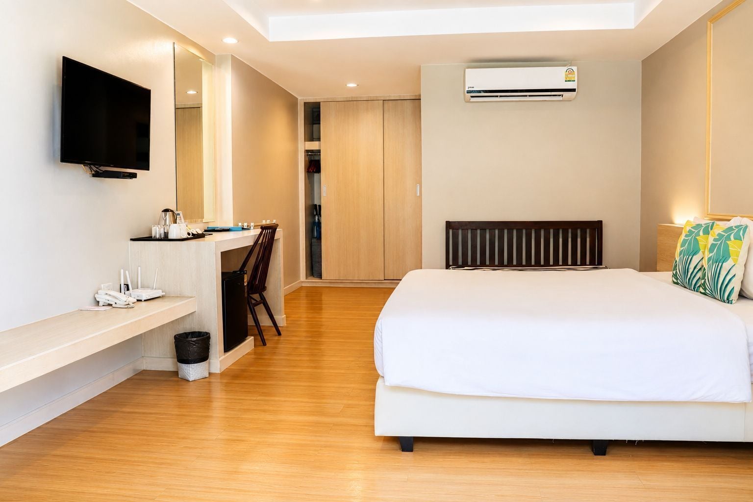 A modern hotel room with a bed, a desk with a chair, a TV, and an air conditioning unit.