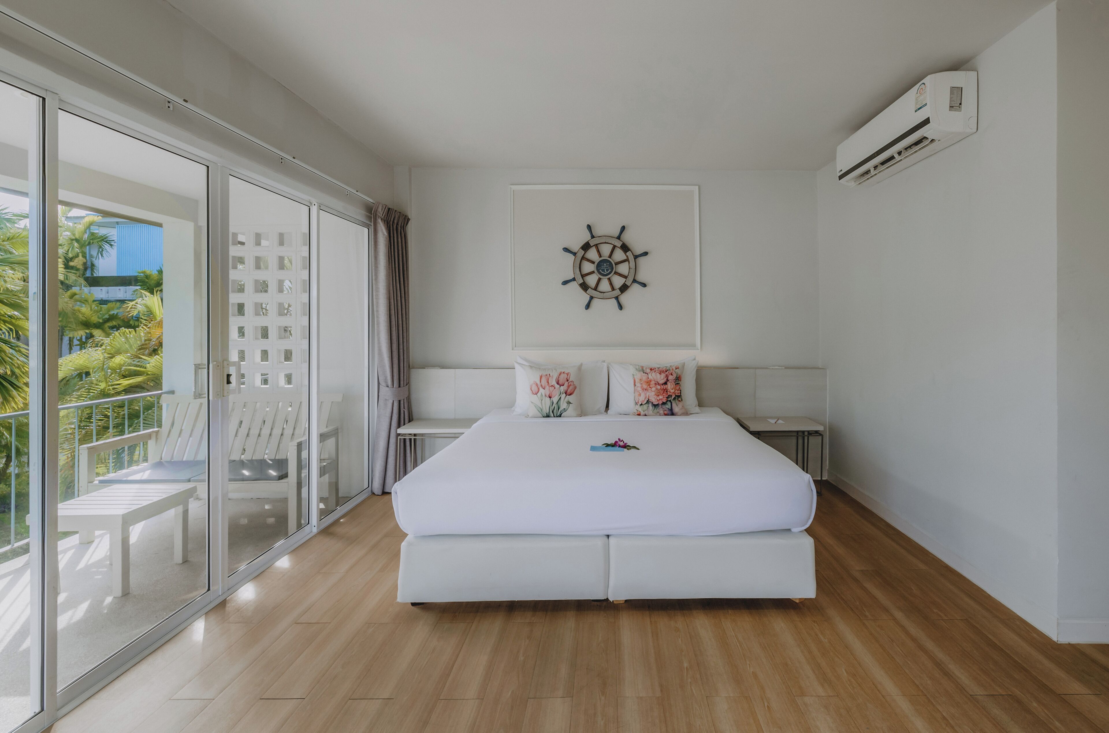 A bedroom with a bed, a wall-mounted air conditioner, a large window with a view of palm trees, and a decorative wall piece.