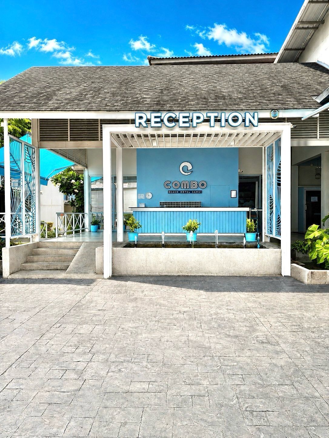 A reception area with a blue wall, a sign reading 'COMBO BEACH HOTEL SAMUI,' and a set of stairs leading up to it.