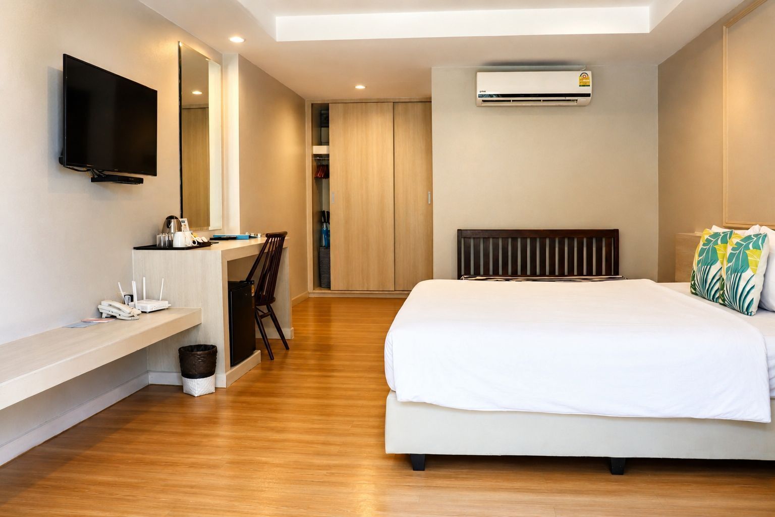 A hotel room with a bed, a desk with a chair, a TV mounted on the wall, and an air conditioning unit.