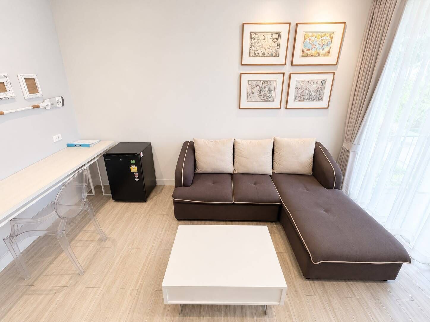 A modern living room with a sectional sofa, a white coffee table, a small black refrigerator, a desk with a laptop, and framed artwork on the wall.