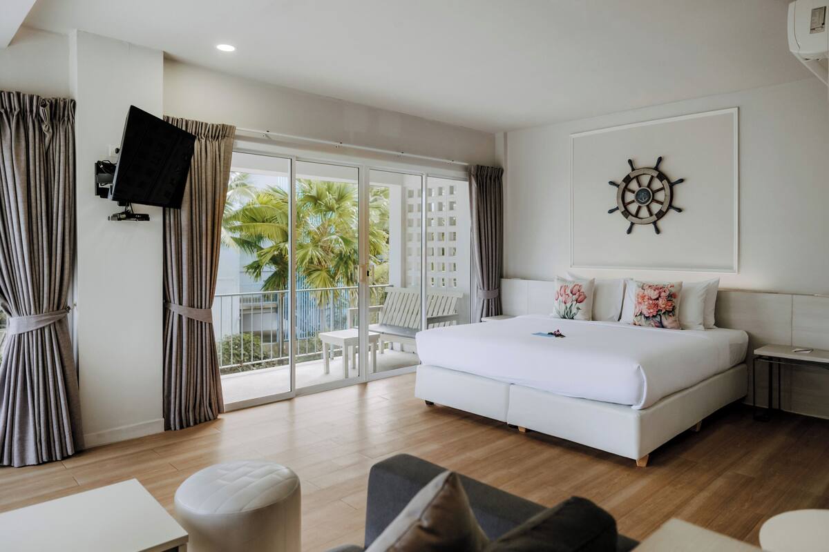 A modern hotel room with a large bed, a flat-screen TV mounted on the wall, a sail wheel decoration, and a balcony with a view of palm trees.