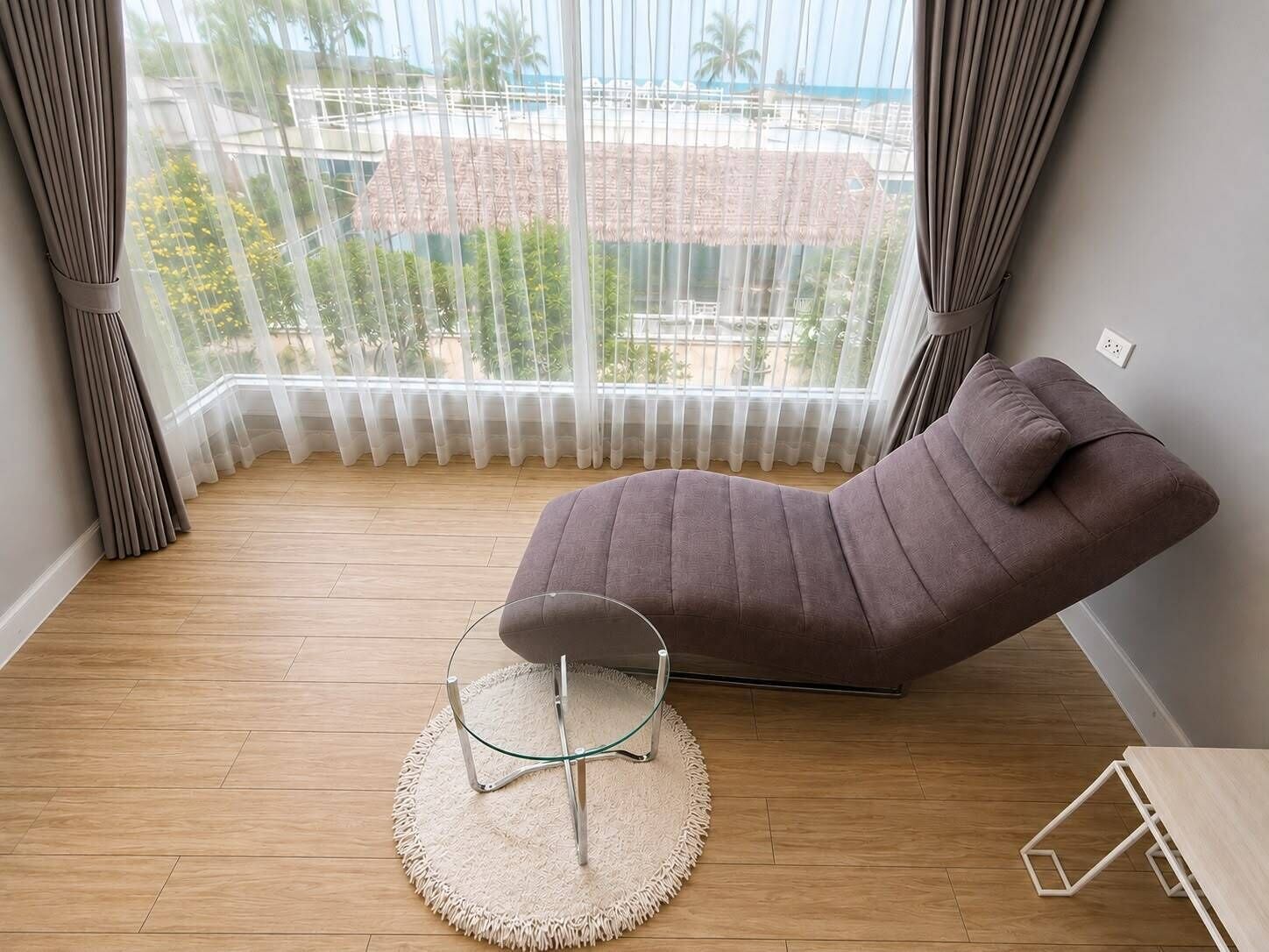 A modern room with a cushioned chair, a glass table, and a view of a beach and palm trees through sheer curtains.