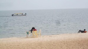 A beach with a rowboat in the water, a chair and a person sitting on the sand, and another person lying down.