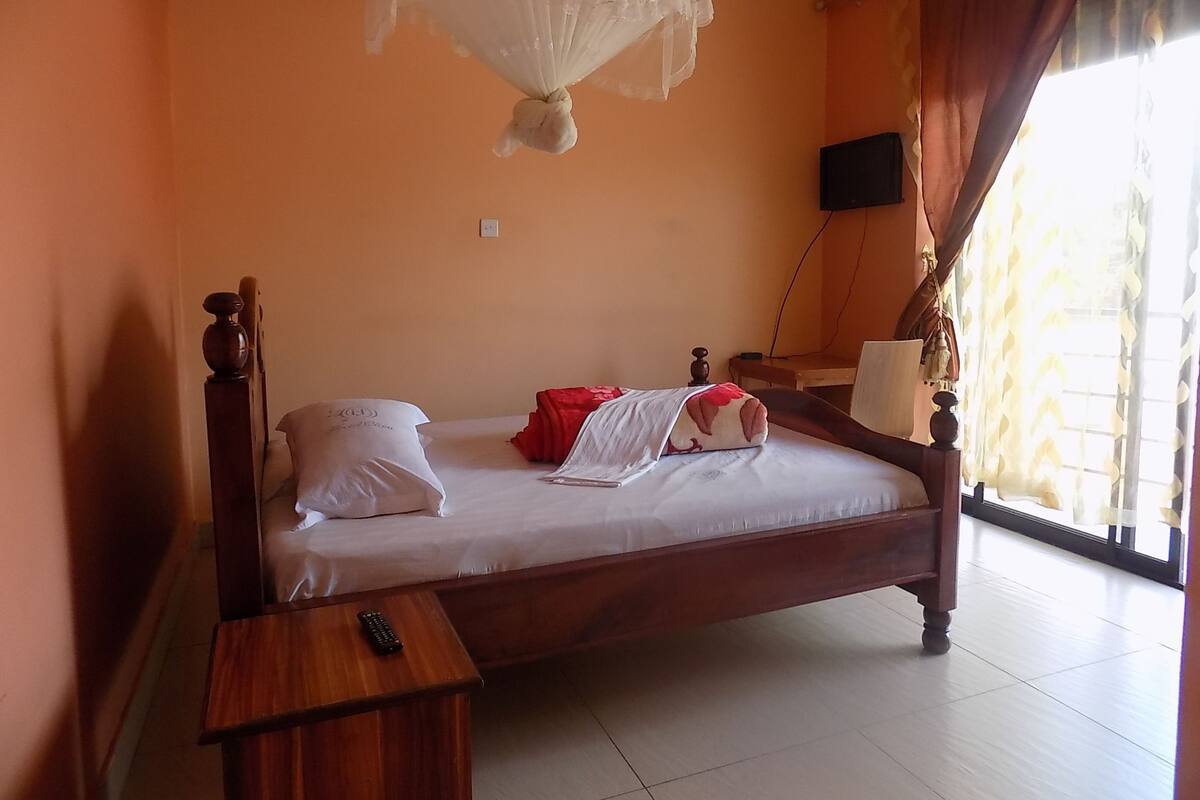 Un lit simple avec un cadre en bois, une table de chevet avec une télécommande, un couvre-lit blanc et un téléviseur fixé au mur.