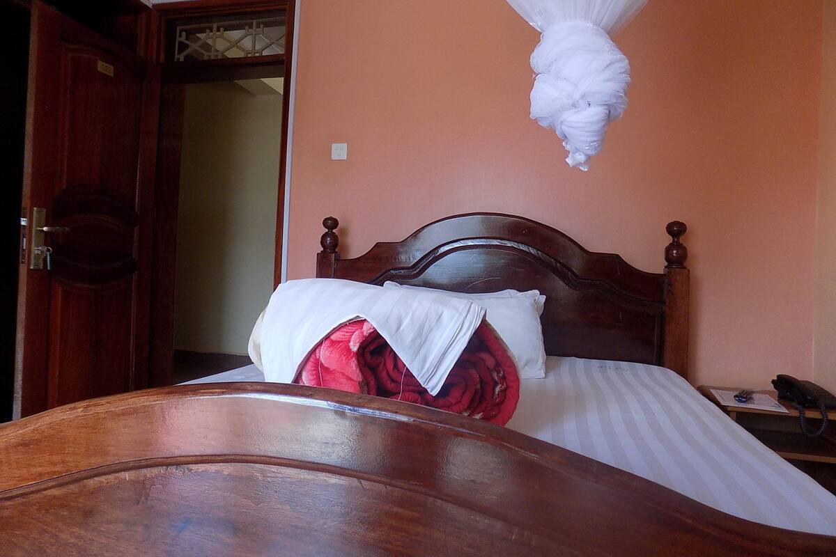 Un lit en bois recouvert d’une courtepointe blanche et orné d’un coussin rouge, une table de chevet avec un téléphone, et une porte avec une plaque numérotée.