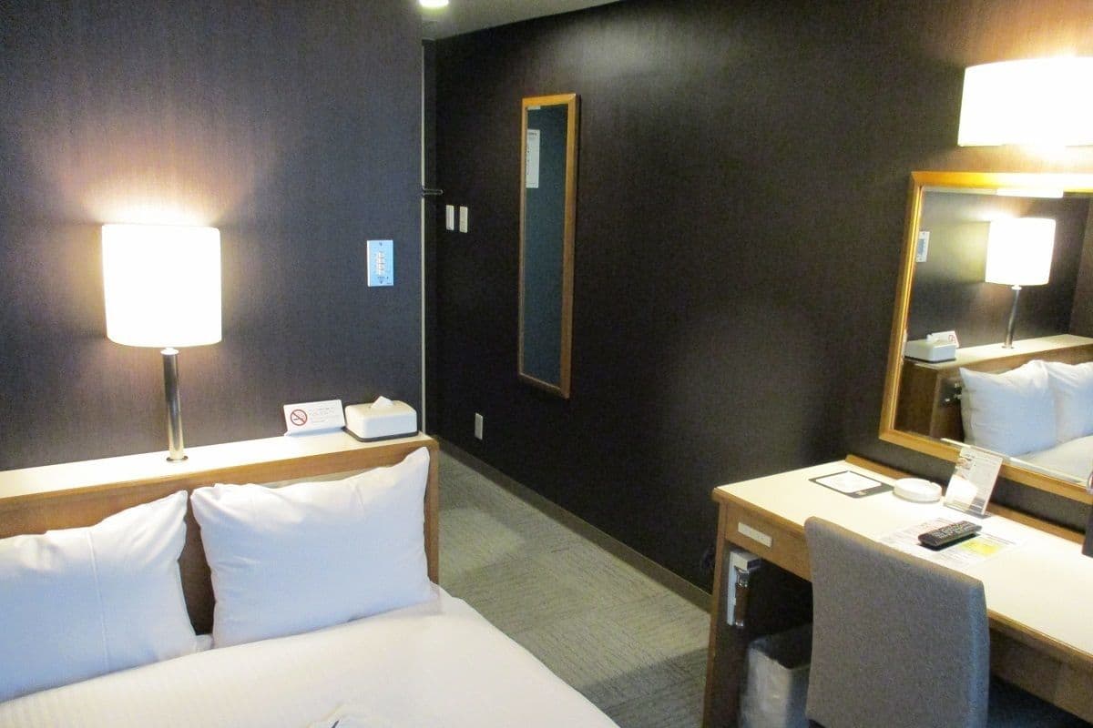 A hotel room with a bed, bedside table, chair, desk, lamp, mirror, and a small trash bin.