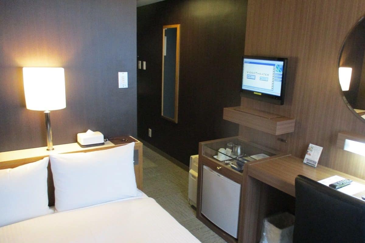 A hotel room with a bed, bedside lamp, desk, chair, TV, and a small refrigerator.