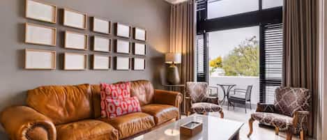 A modern living room with a leather sofa, a coffee table, patterned armchairs, a dining table, and a large window with blinds.