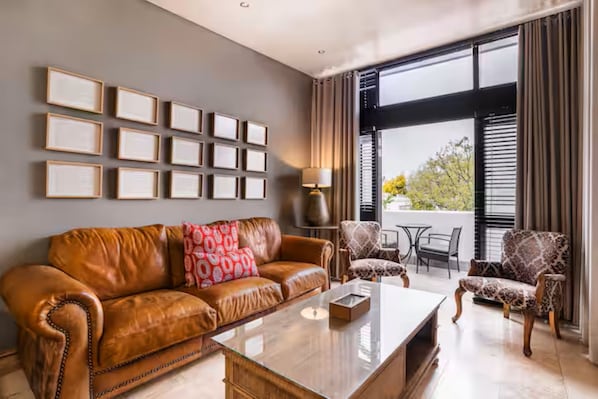 A modern living room with a leather sofa, a coffee table, patterned armchairs, a dining table, and a large window with blinds.