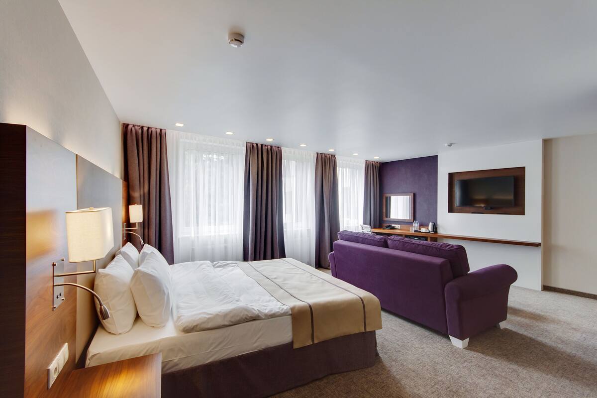 A hotel room with a bed, a sofa, a TV mounted on the wall, and a window with curtains.