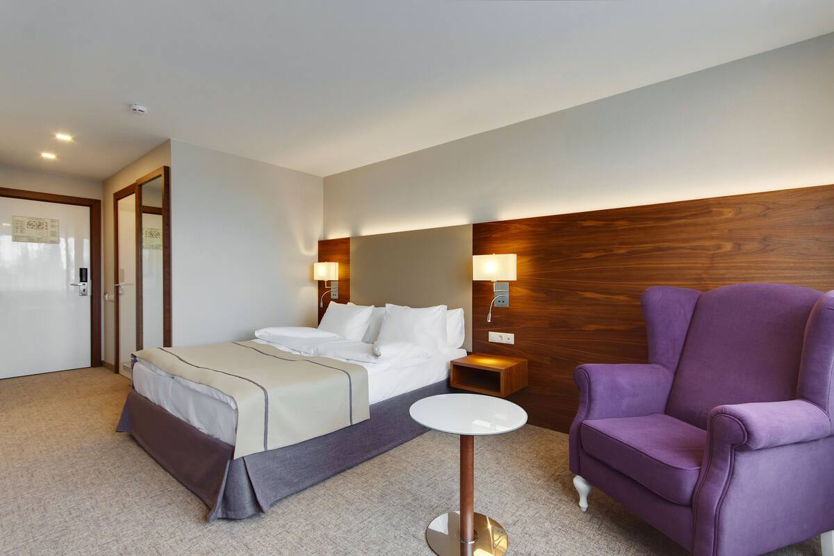 A hotel room with a bed, a purple armchair, a small table, and a wooden headboard.