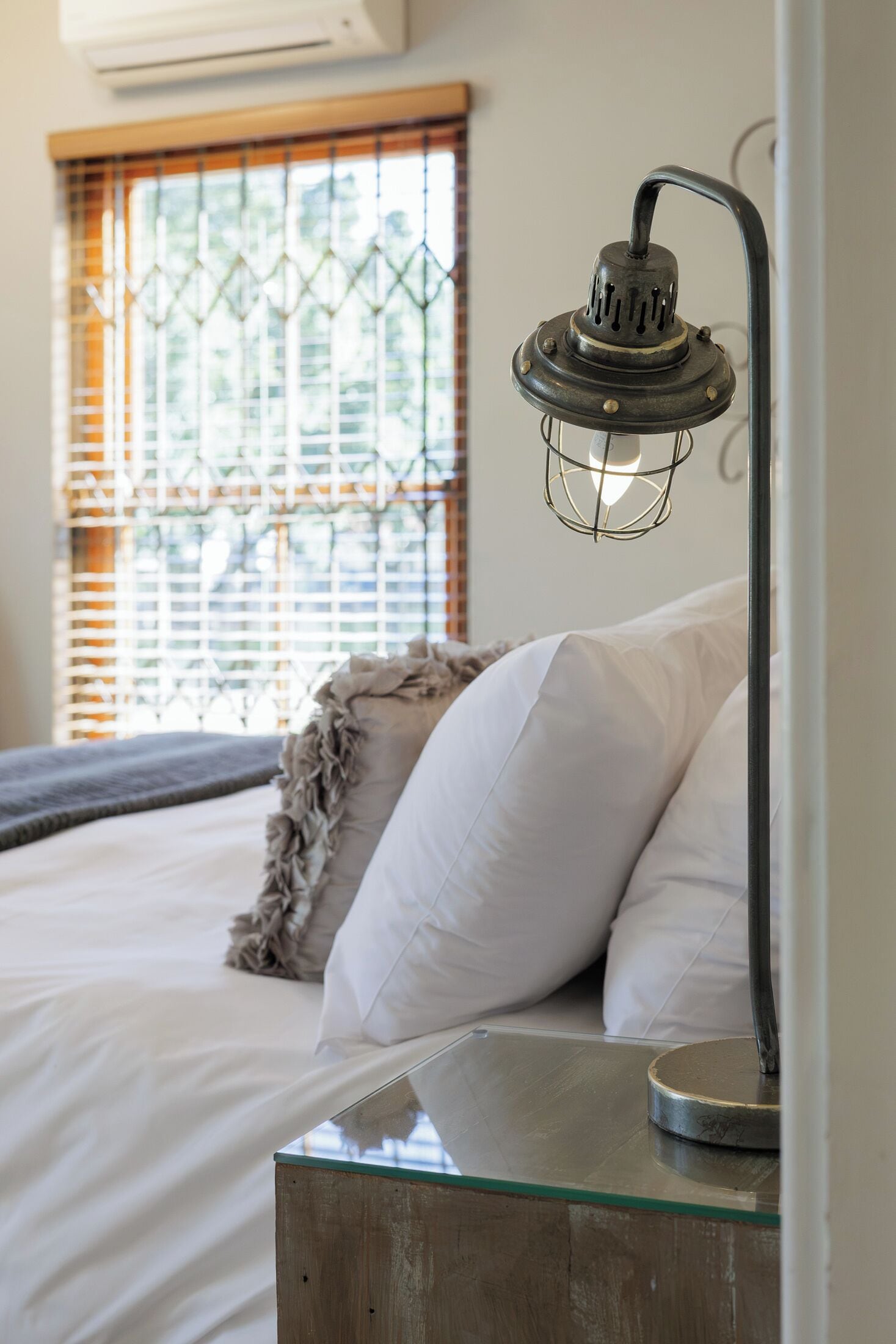 A bed with white linens and a decorative pillow, a glass table with a metal lamp, and a window with blinds.