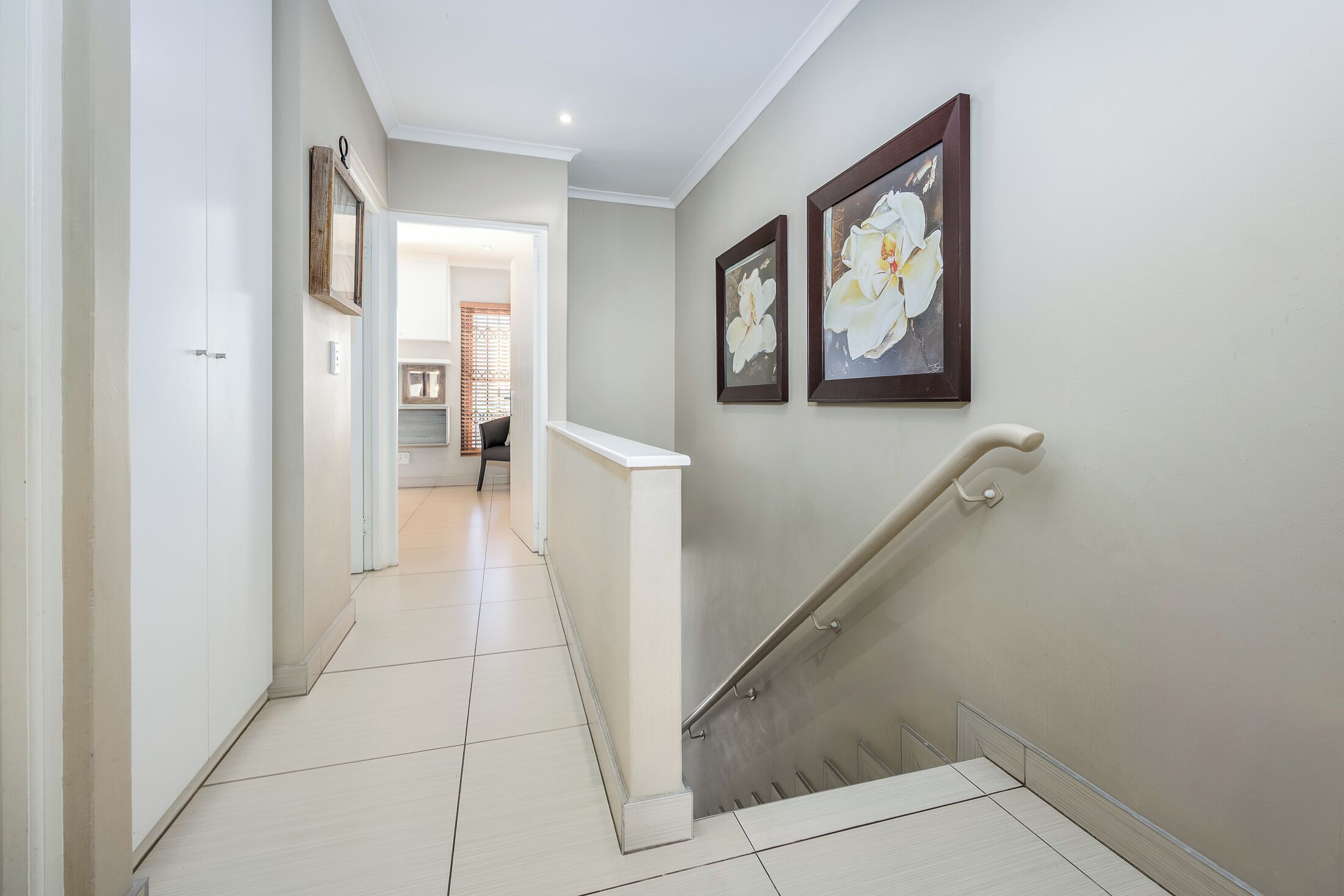 A hallway with a staircase, white walls, and two framed floral paintings.
