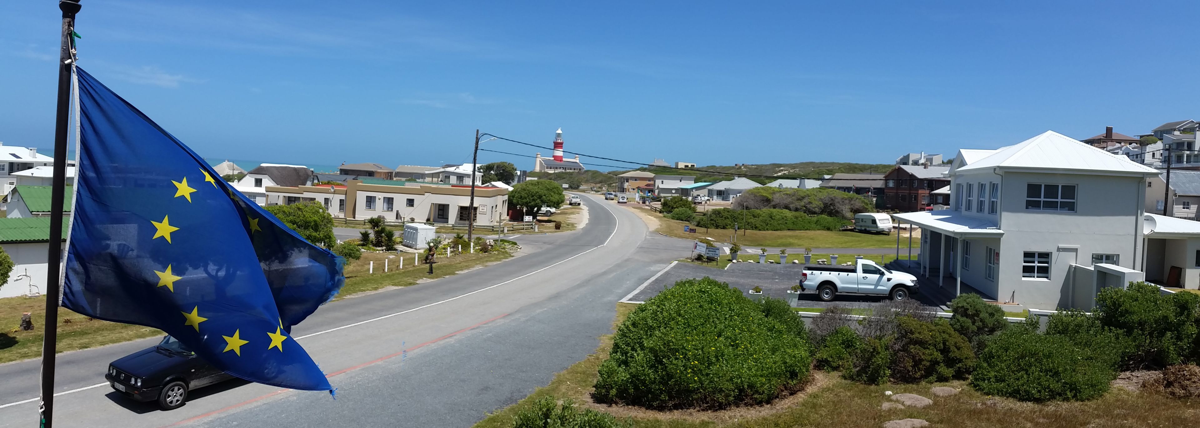 A coastal town with a prominent lighthouse, a road curving through the center, and a flagpole with the European Union flag.