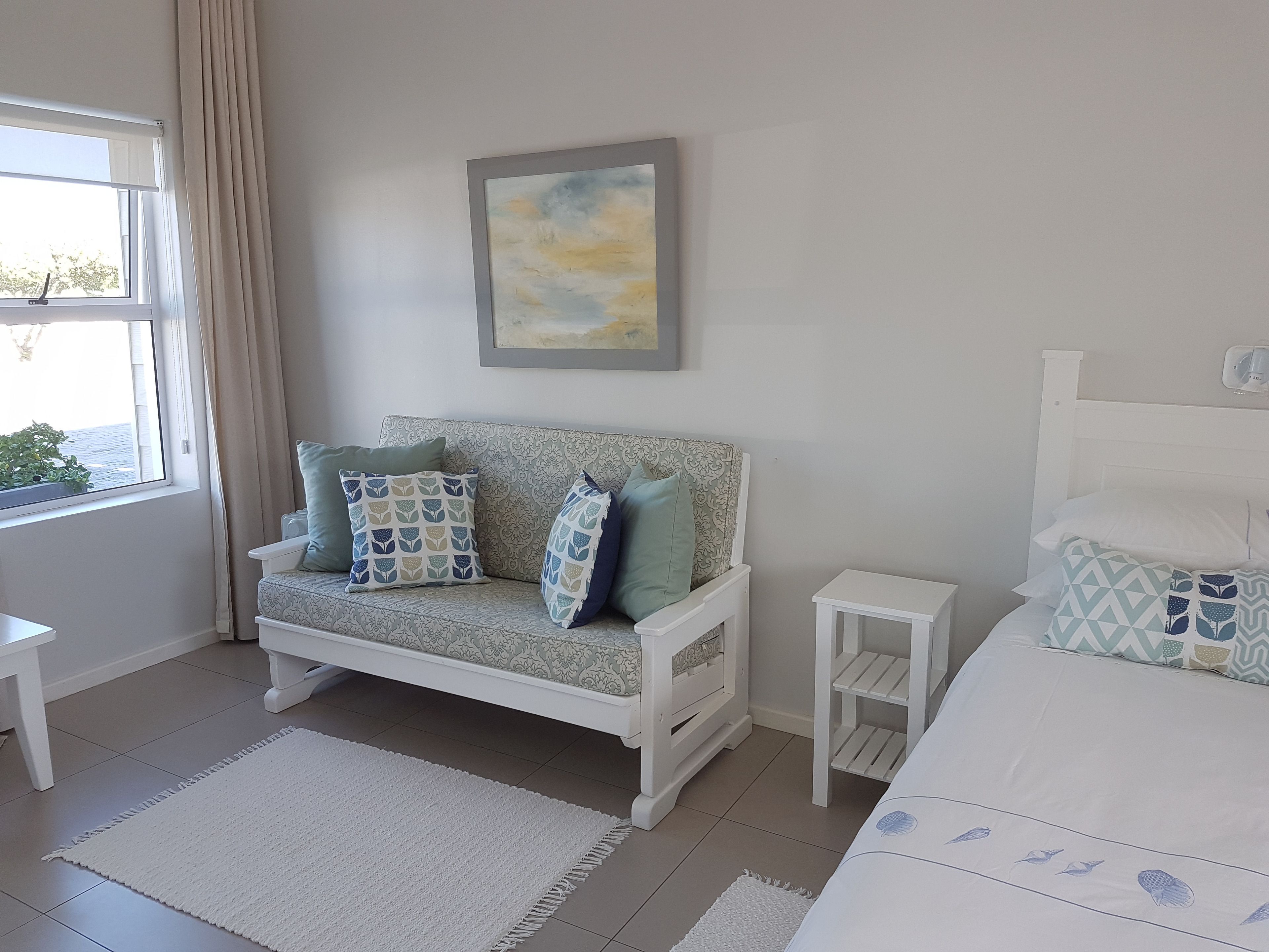 A bedroom with a sofa bed, a bedside table, a small bedside table, a window with curtains, and a painting on the wall.