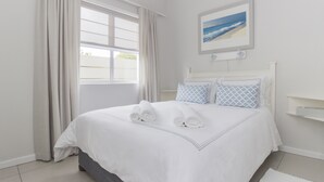 A neatly made bed with white linens and rolled towels, a window with curtains, and a framed picture on the wall.