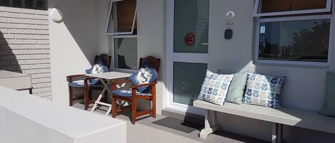 Un balcón con una mesa y sillas blancas, un banco con cojines estampados y una ventana con un cartel de 'no fumar'.