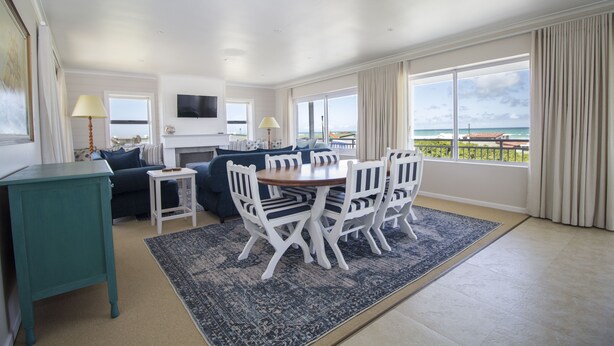 A spacious living room with a dining area, large windows with a view of the ocean, a flat-screen TV mounted on the wall, and a teal side table.