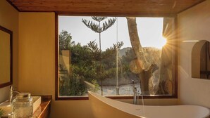 Un baño moderno con una bañera independiente, un ventanal con vistas a un paisaje natural y un tocador de madera con un tarro de vidrio.
