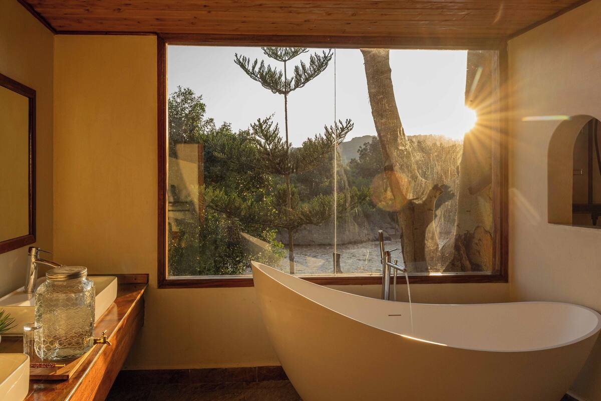 Une salle de bain moderne avec une baignoire sur pieds, une grande fenêtre donnant sur un paysage naturel et un meuble-lavabo en bois surmonté d’un pot en verre.