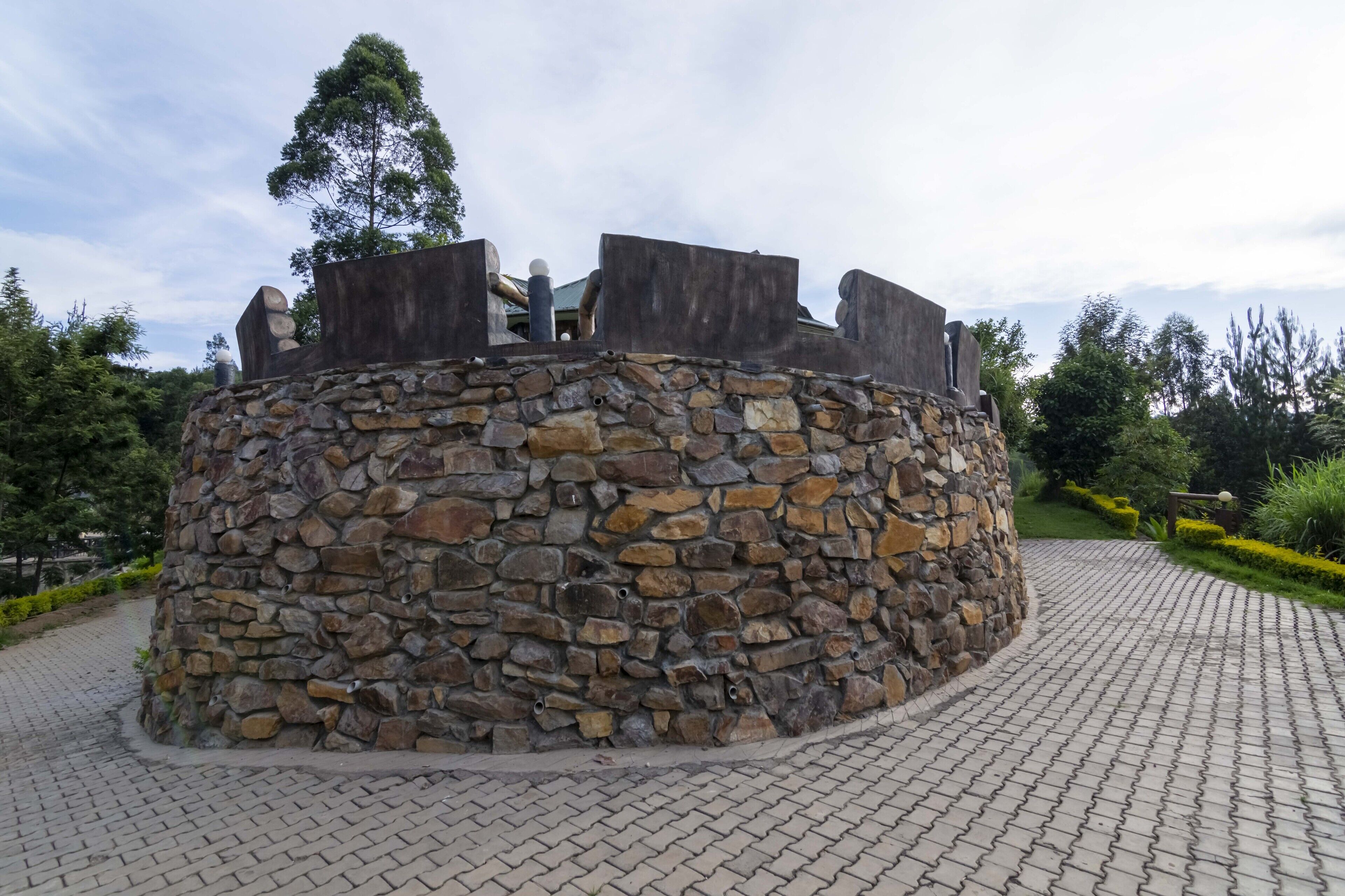 Estrutura de pedra com base circular e topo plano, rodeada por um caminho pavimentado e vegetação.