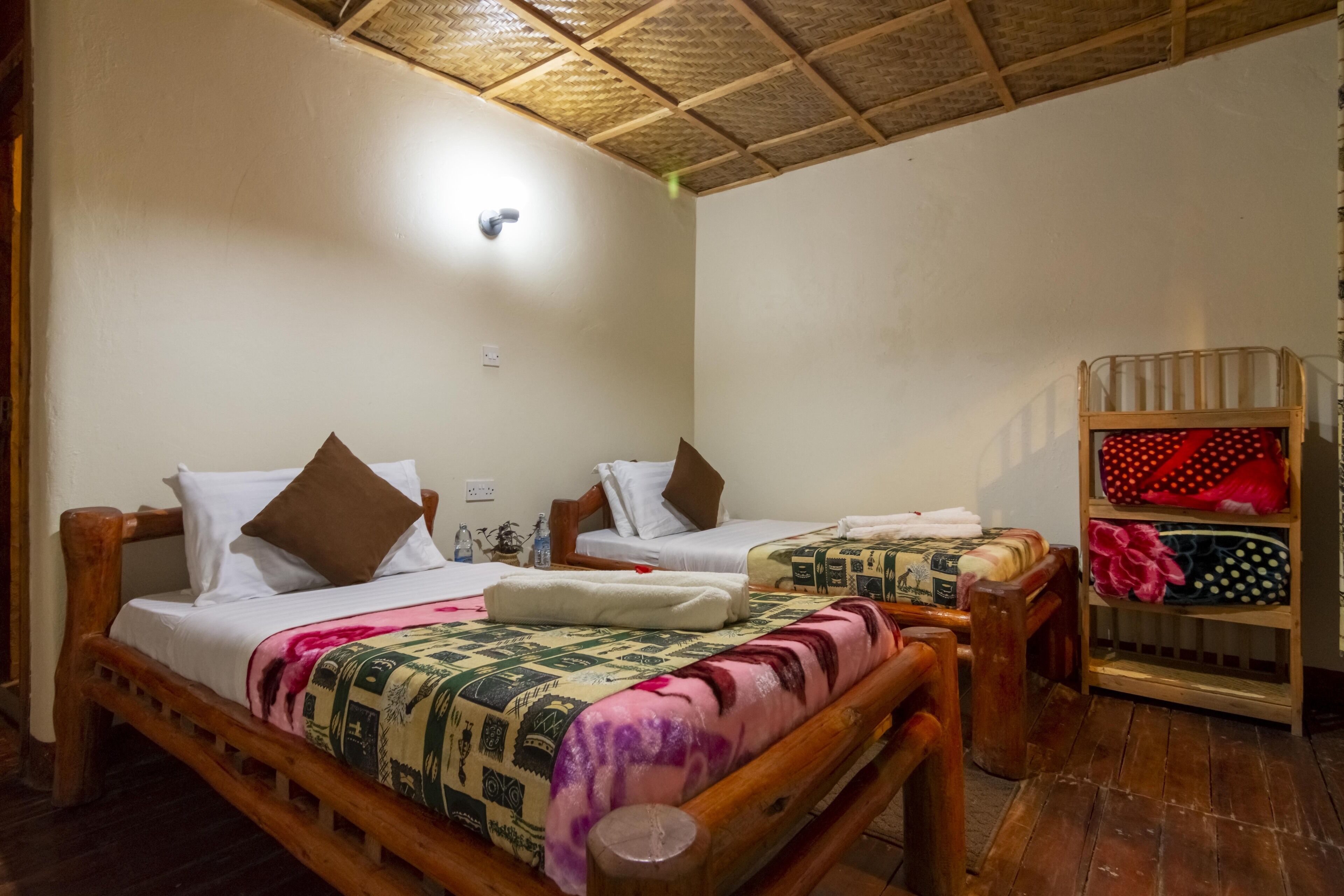 A room with two wooden beds, each with a pillow and a blanket. There is a shelf with folded clothes and a small table with a lamp.