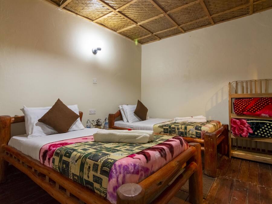 A room with two wooden beds, each with a pillow and a blanket. There is a shelf with folded clothes and a small table with a lamp.