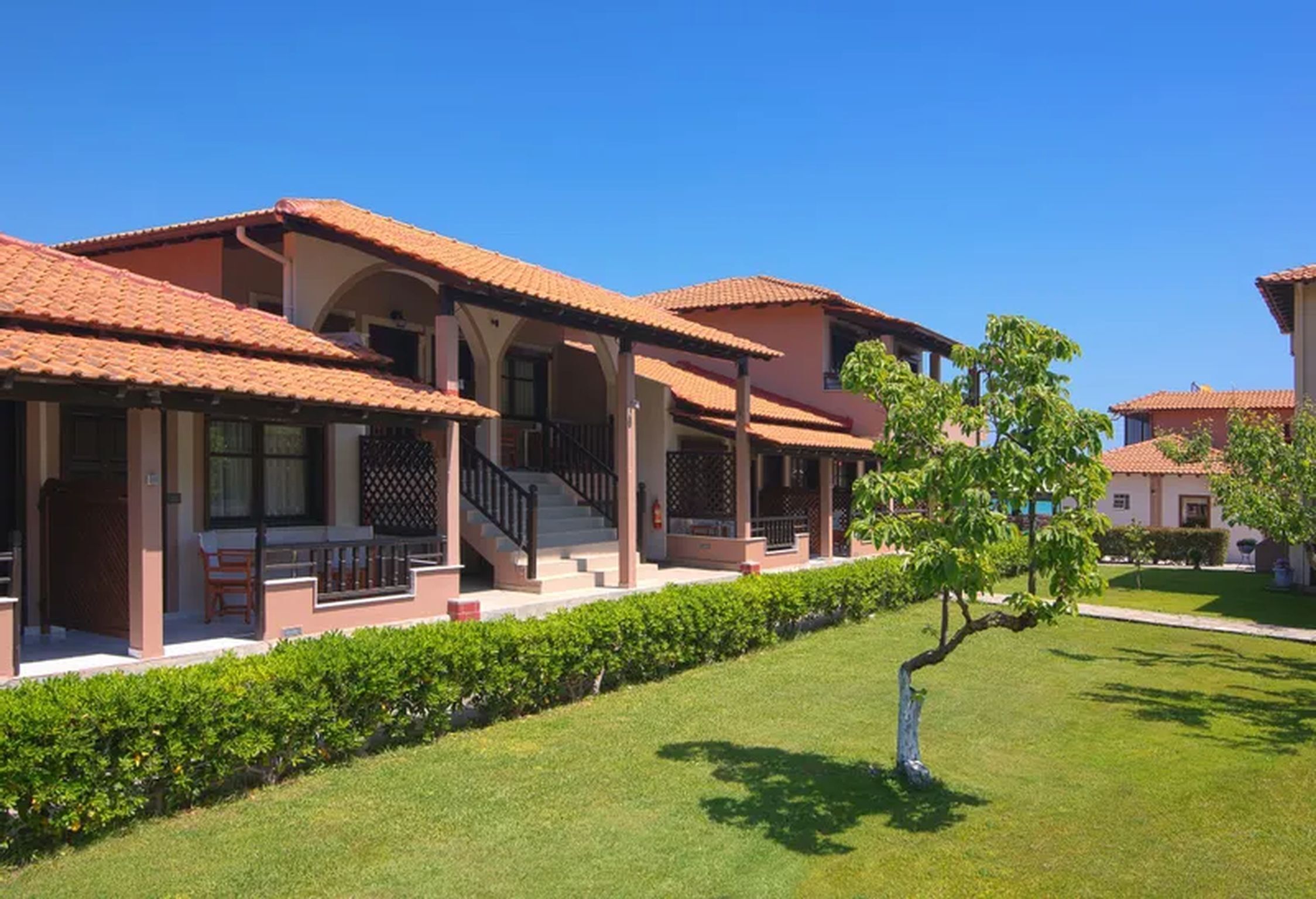 A row of modern buildings with red-tiled roofs and covered balconies, surrounded by well-maintained lawns and a small tree.