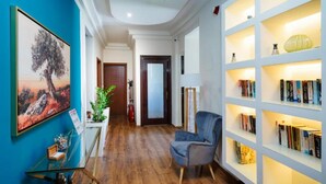 A hallway with a blue wall, a glass table with a plant, a blue armchair, and a bookshelf with books. - Kamelia & Semeli Hotel (Thasos)