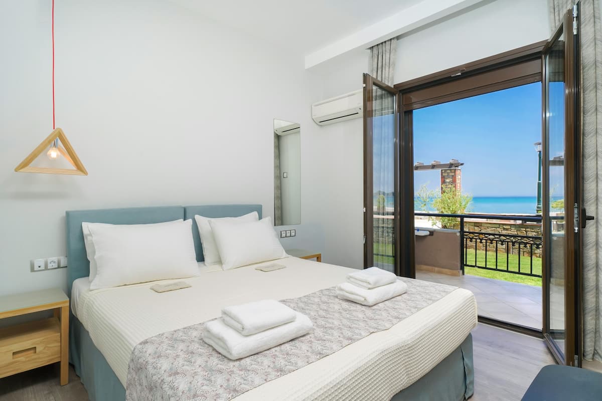 Un dormitorio con una cama grande, una mesita de noche, una lámpara colgante y vista al mar desde una puerta abierta.