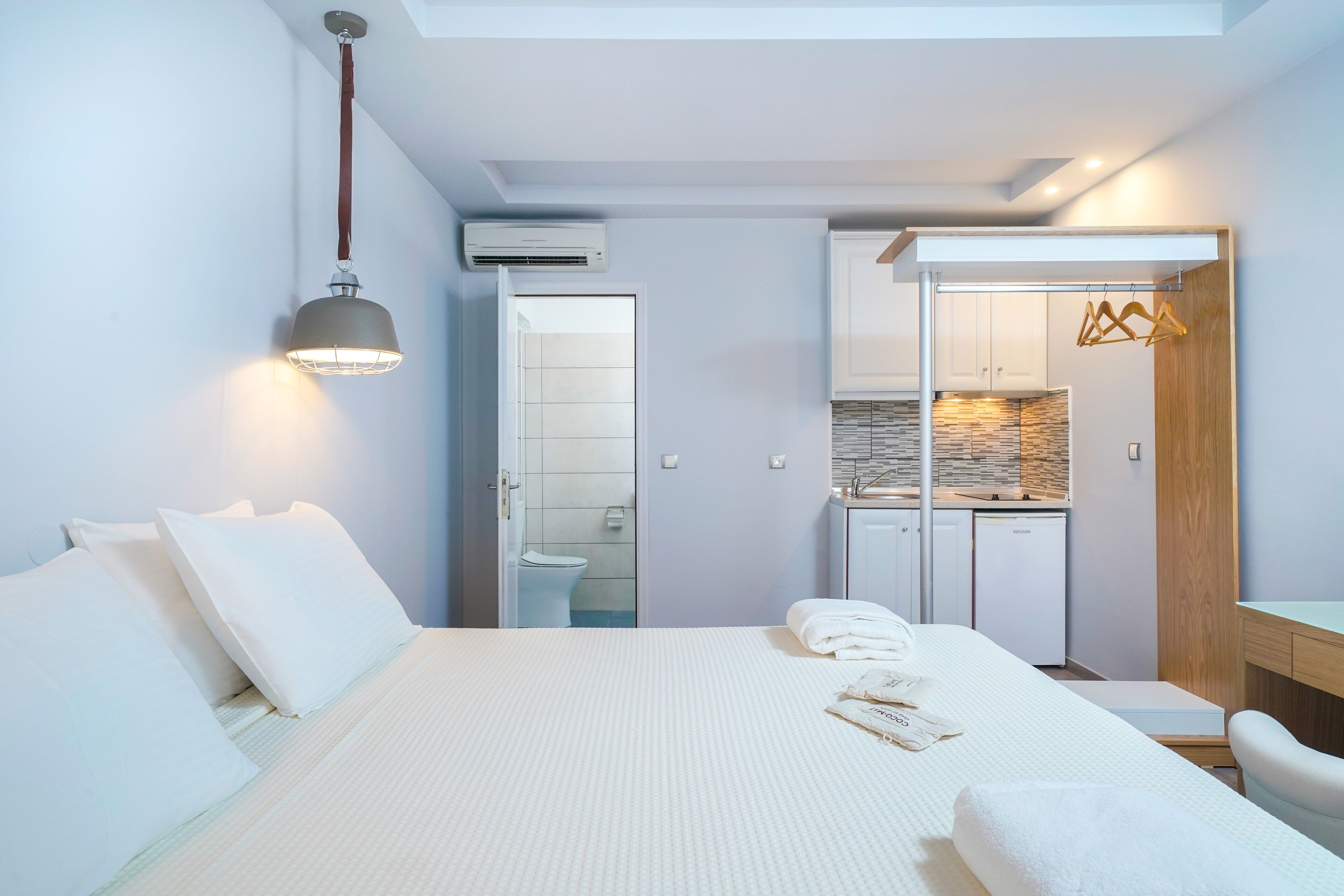 A modern hotel room with a large bed, a bedside table, a desk, and a kitchenette.