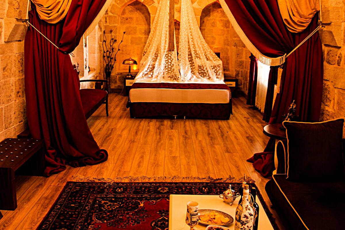 A room with a bed under a canopy, a chandelier, a table with fruit and a bottle, and a red and yellow draped window.