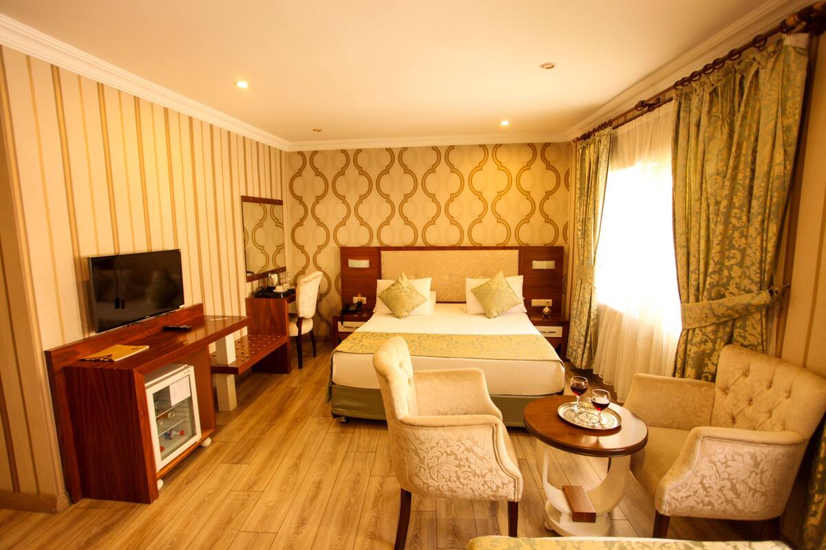 A hotel room with a large bed, a desk with a TV, two armchairs, and a small table with a tray.