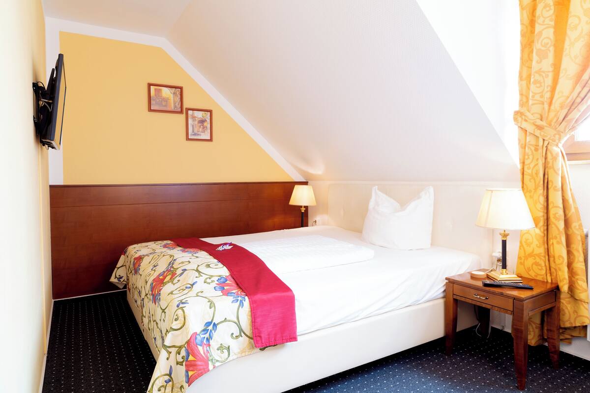 Een hotelkamer met een bed, twee nachtkastjes, een wandtelevisie en een raam met gordijnen.