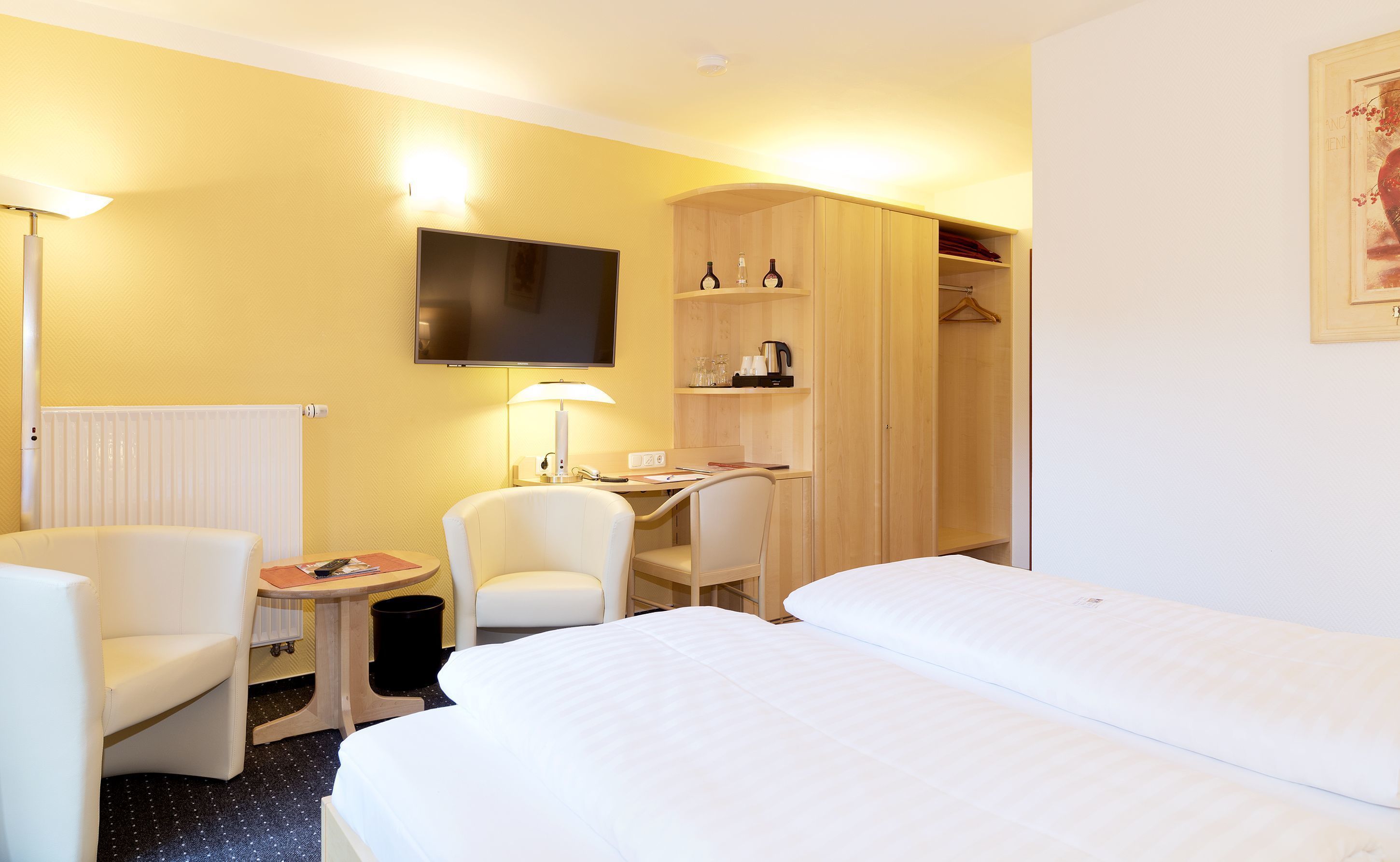 Quarto de hotel com cama, duas cadeiras, escrivaninha, televisão e guarda-roupa.