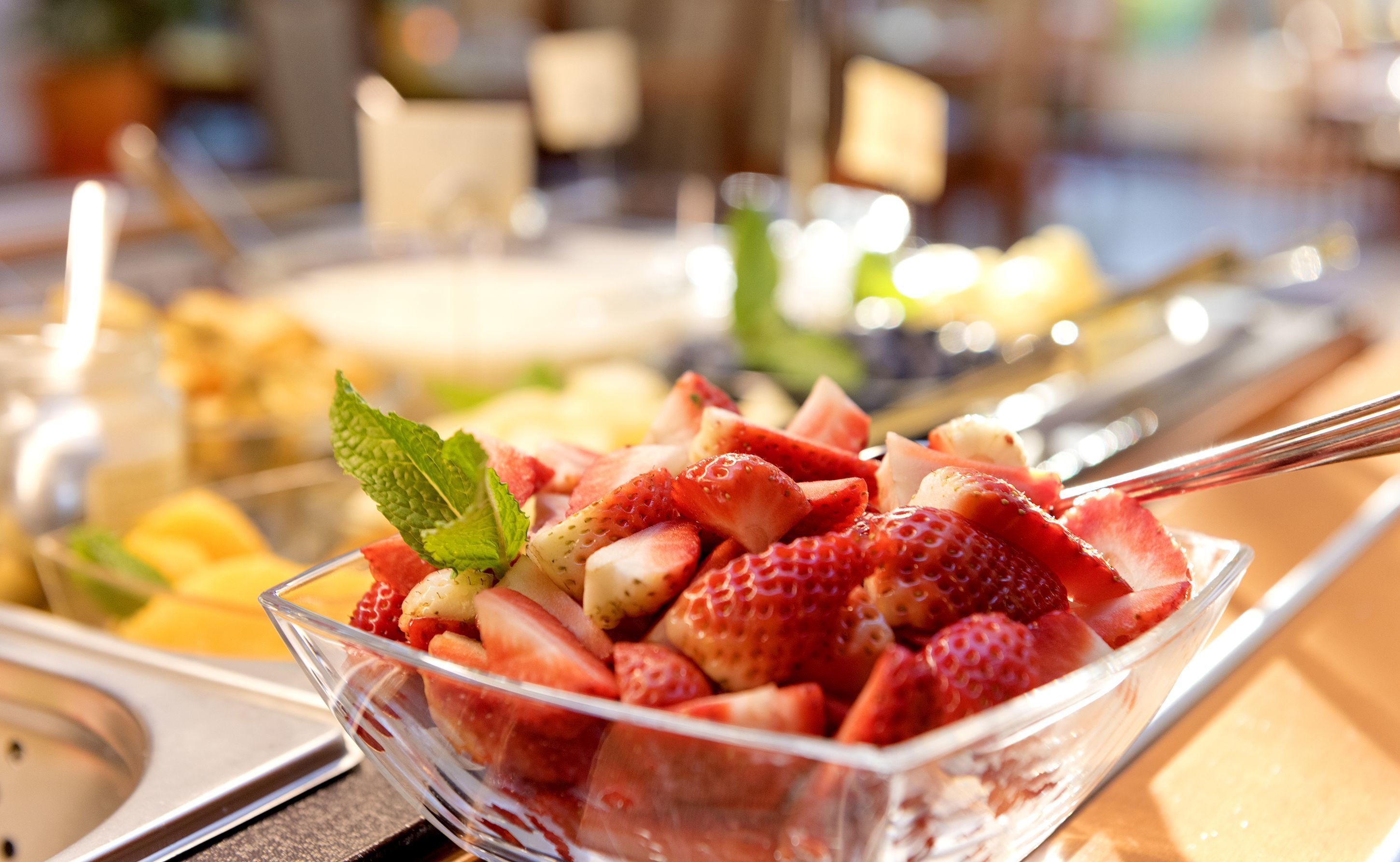 A clear glass bowl filled with sliced strawberries and watermelon, garnished with mint leaves, placed on a buffet table.