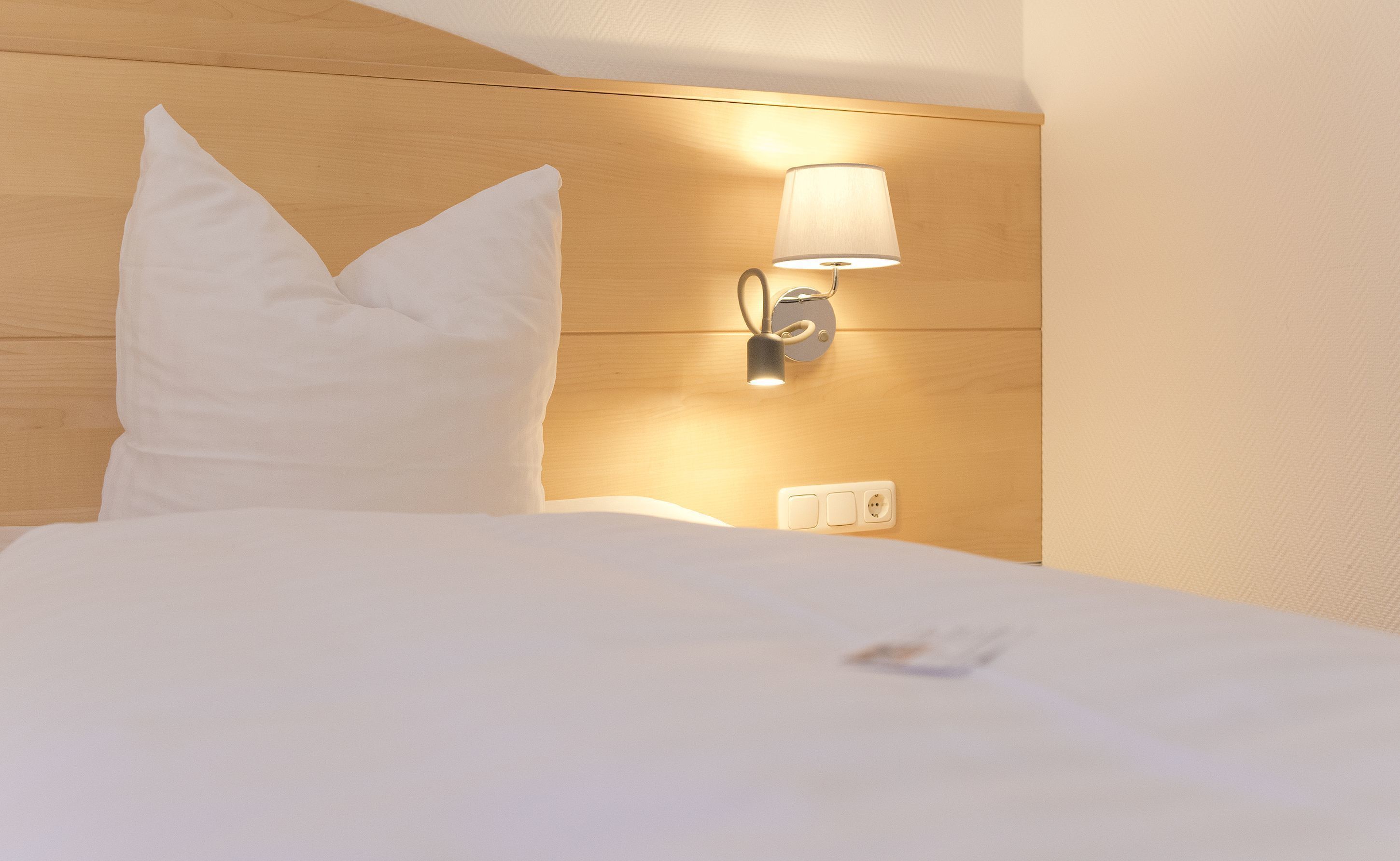 Ein ordentlich bezogenes Bett mit weißer Bettwäsche und Kissen, eine Wandlampe sowie eine Steckdose am Holz-Kopfteil.