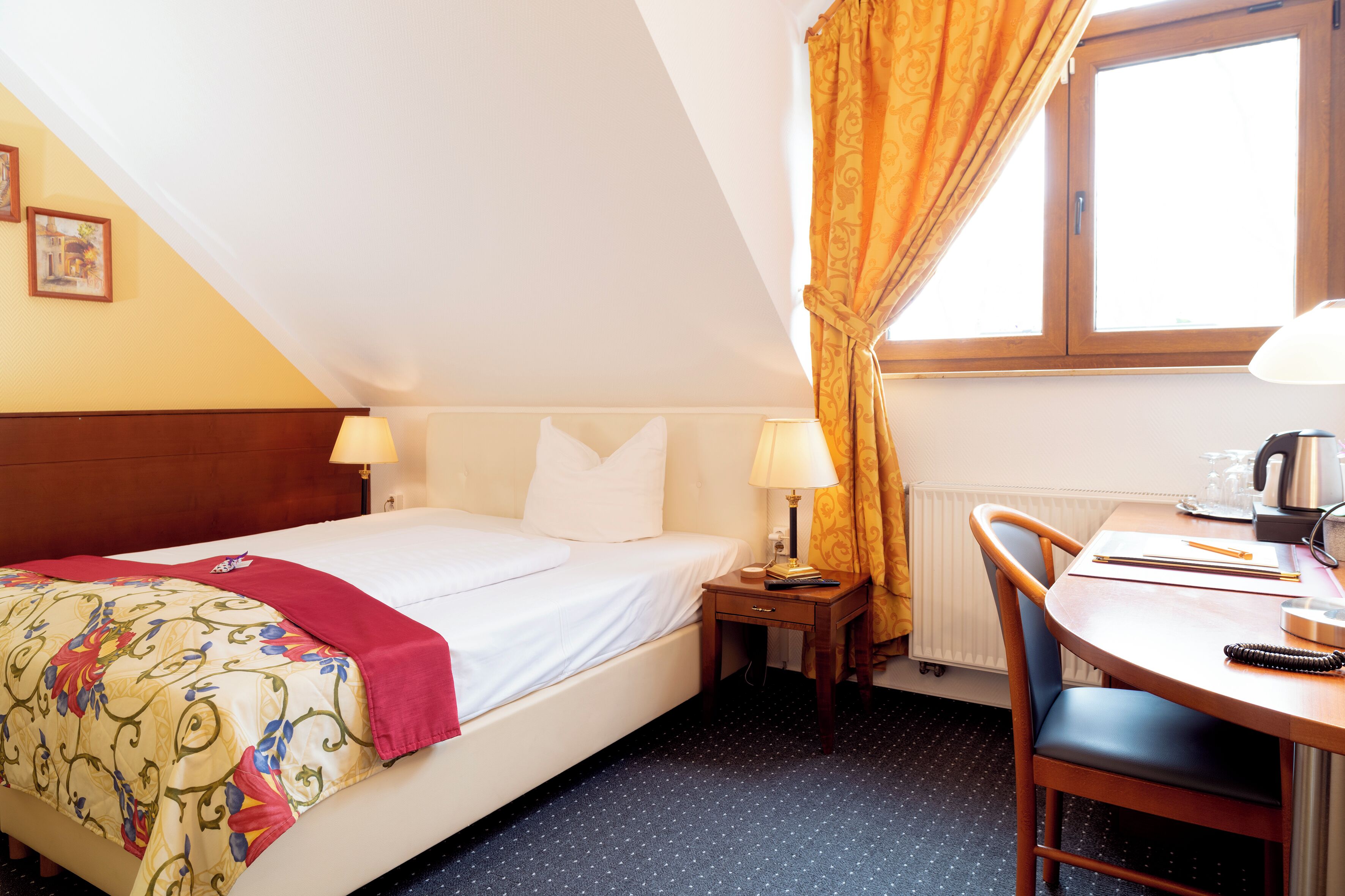 A hotel room with a bed, bedside lamps, a small table with a chair, and a window with curtains.