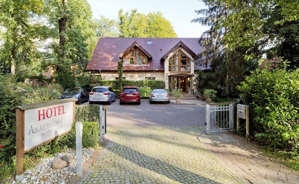 L’entrée d’un hôtel, avec une allée pavée, des voitures garées et une végétation luxuriante.