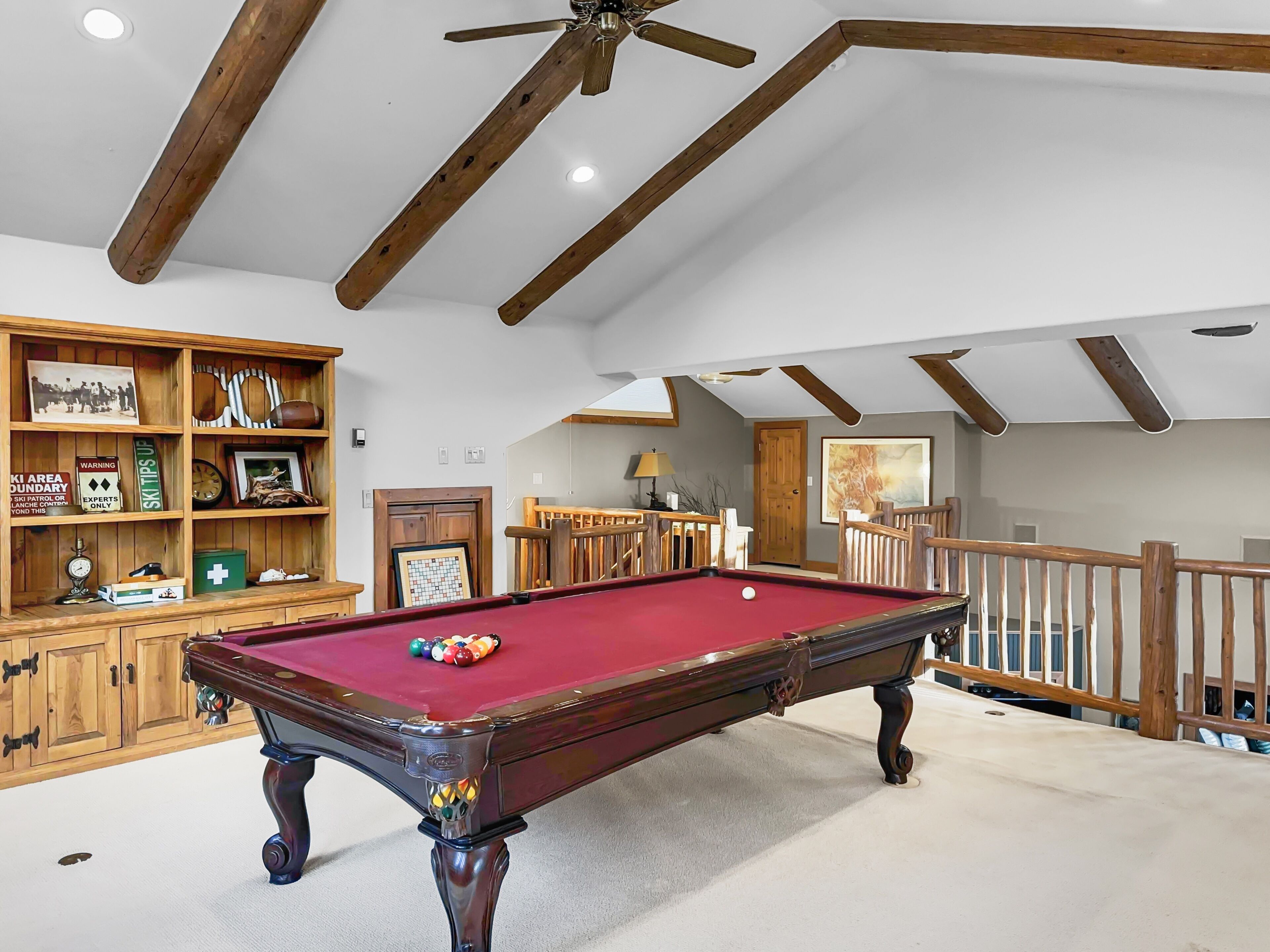 A pool table with a red felt surface, wooden cabinets with various items, and a ceiling fan in a room with wooden beams and a staircase.
