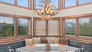A dining area with a wooden table, chairs, and a chandelier. There are three windows with blinds, and a view of greenery outside. - Above the Fray (Breckenridge)
