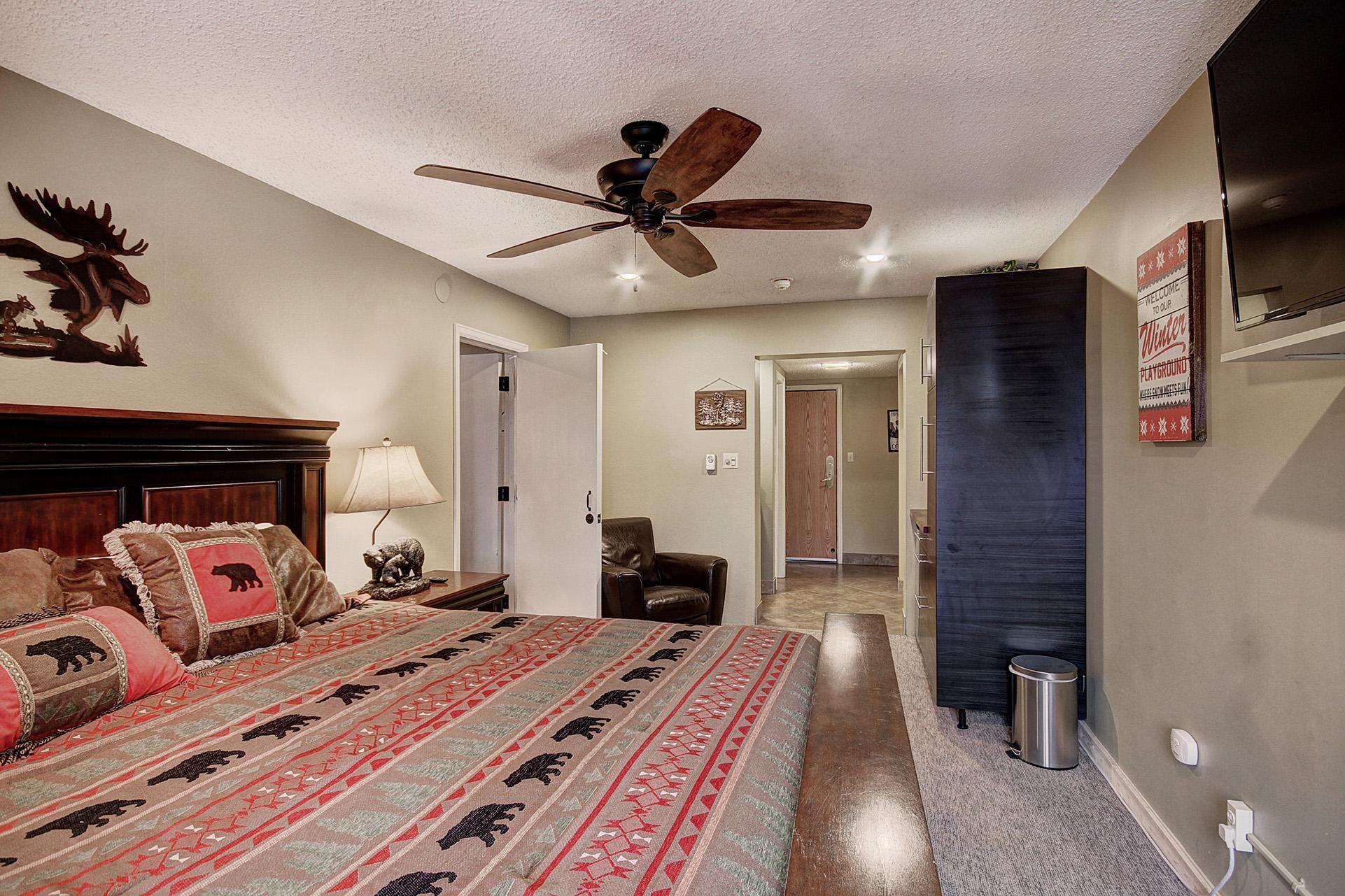 A bedroom with a bed, a ceiling fan, a lamp, a chair, and a door leading to another room.
