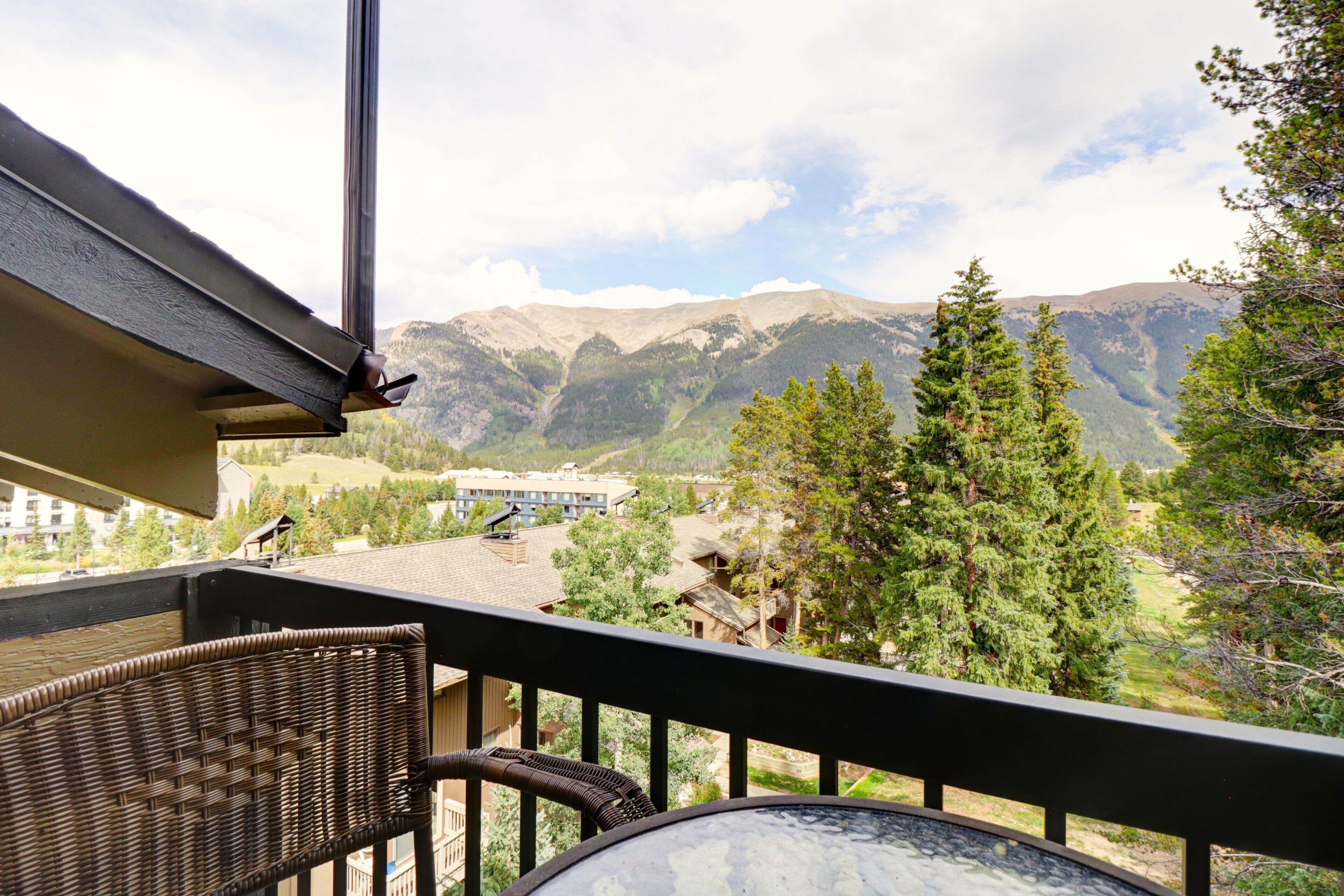 A balcony with wicker furniture, a glass table, and a view of mountains and buildings.