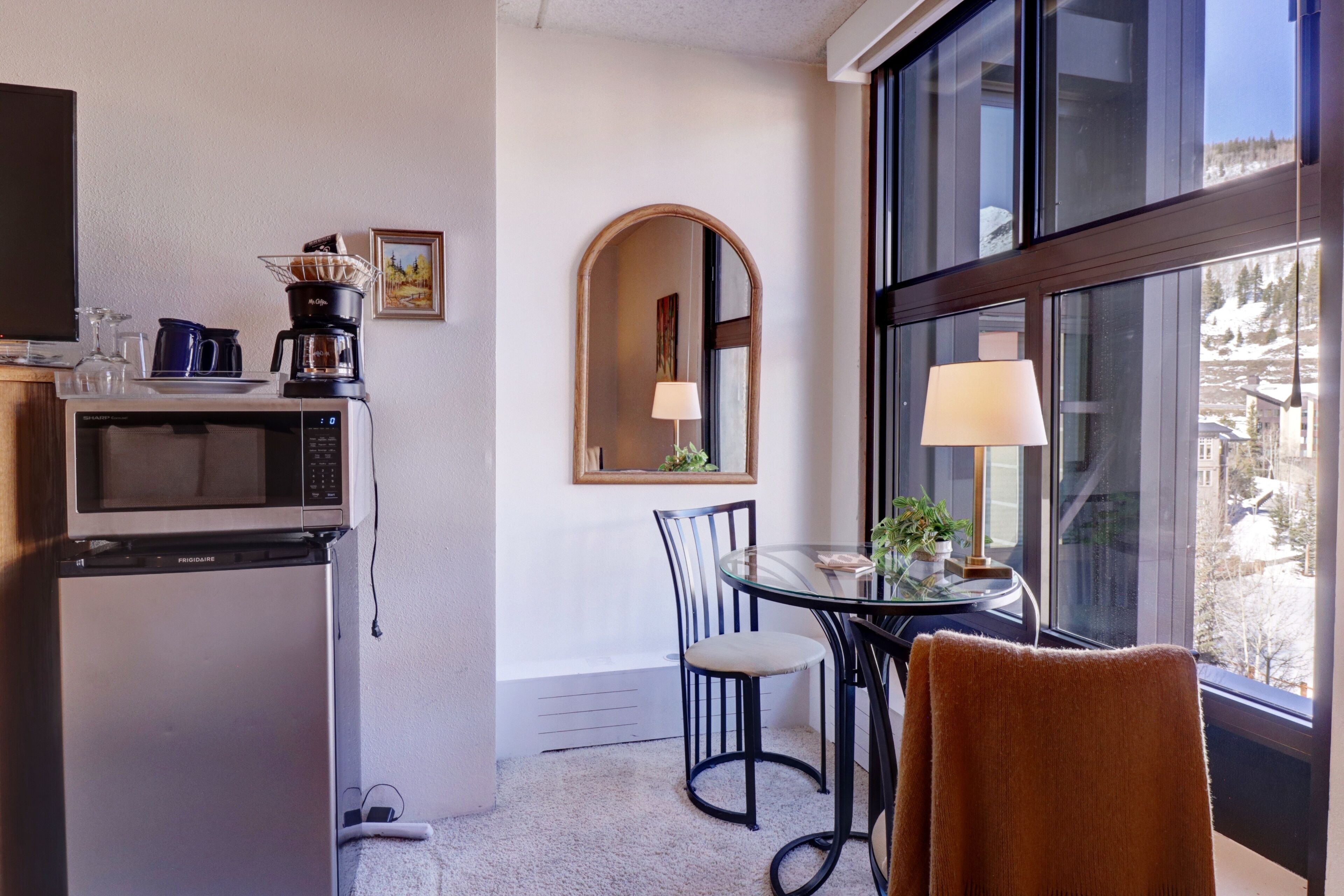 A compact hotel room with a kitchenette, a dining area with a glass table and chairs, a large window with a view of a snowy landscape, and a mirror on the wall.