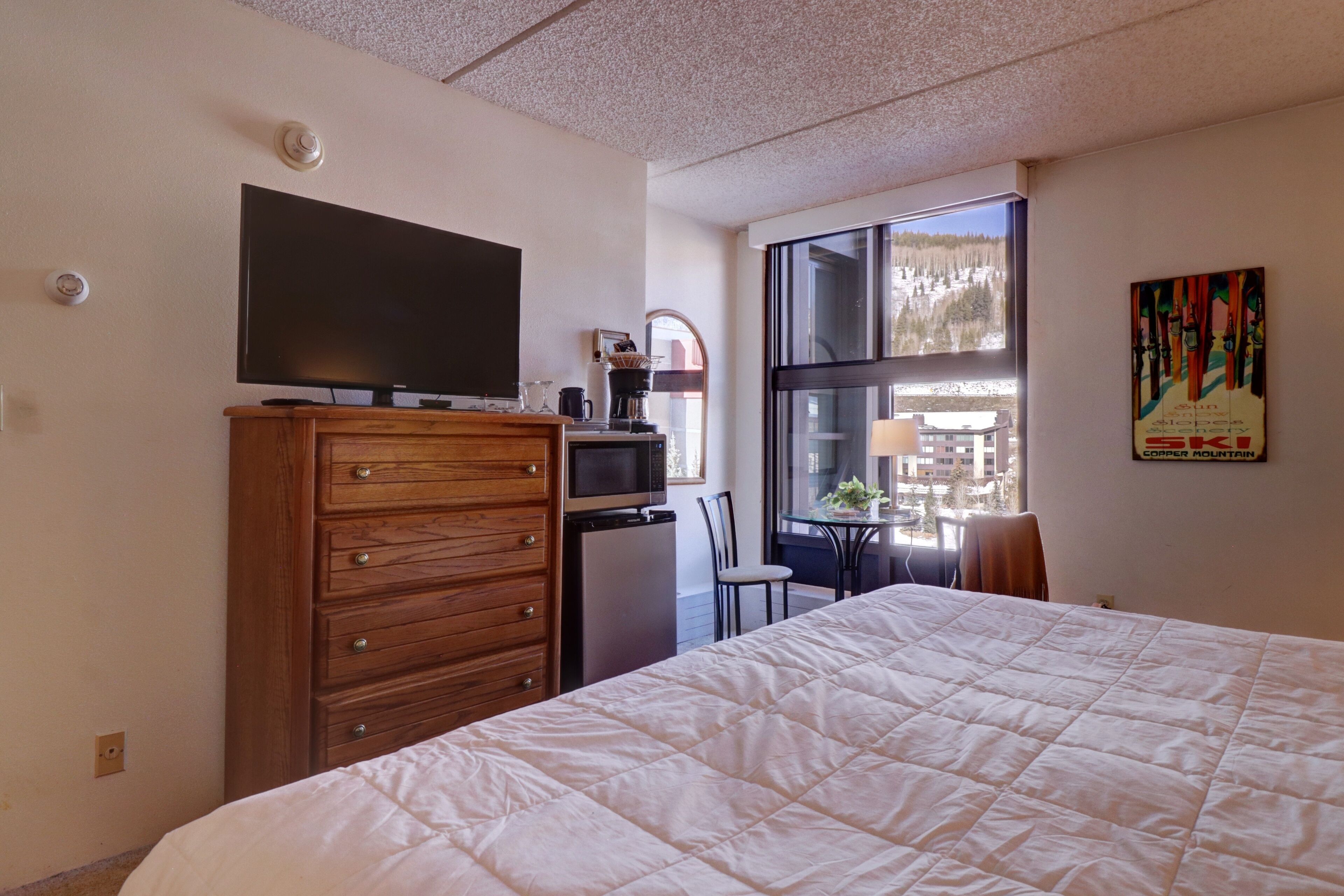 Ein Hotelzimmer mit einem großen Bett, einem hölzernen Schminktisch, einem Flachbildfernseher, einer kleinen Küchenzeile und einem Fenster mit Blick auf eine verschneite Landschaft.