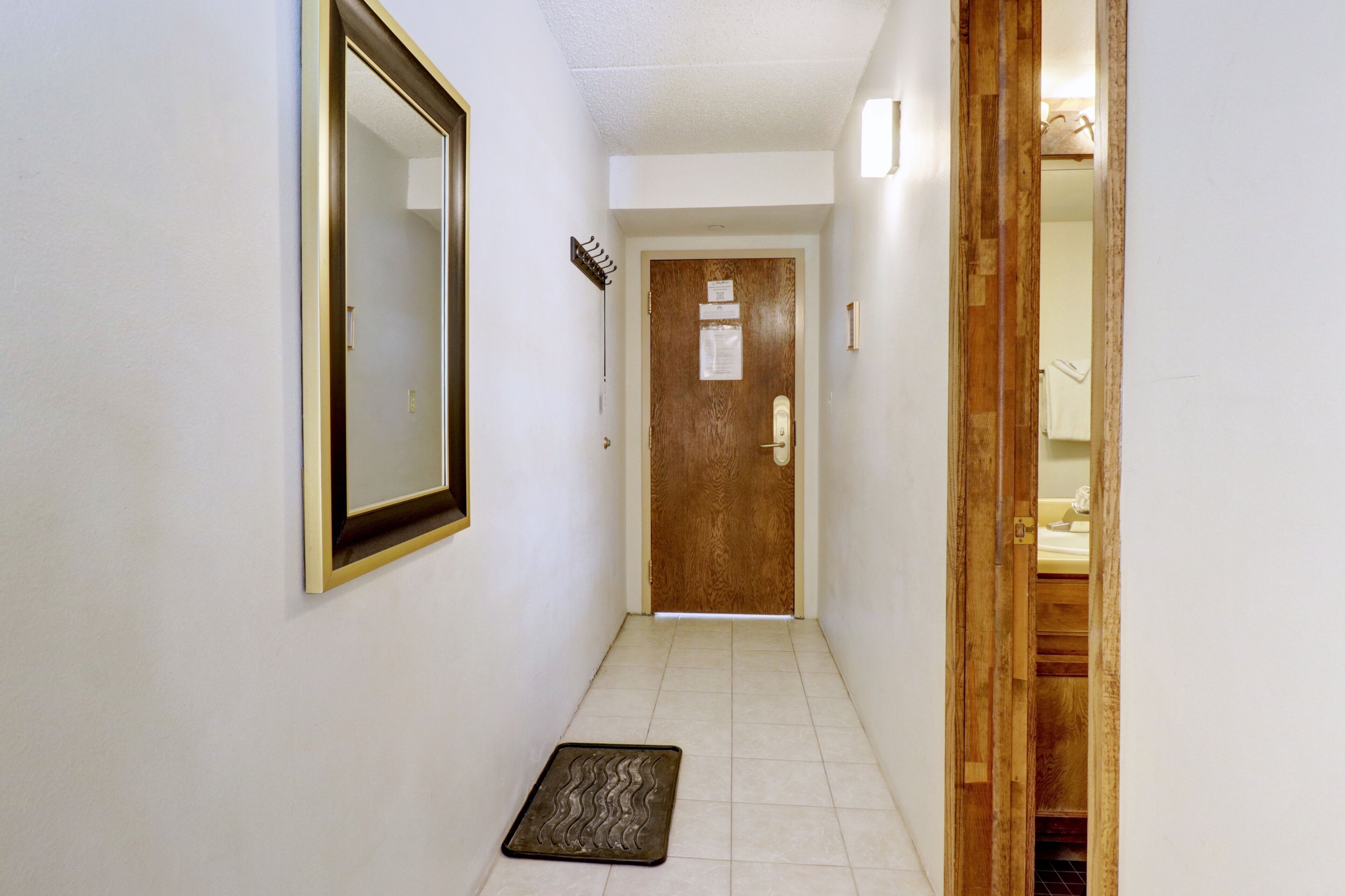 A narrow hallway with a wooden door at the end, a doormat, and a wall-mounted mirror.
