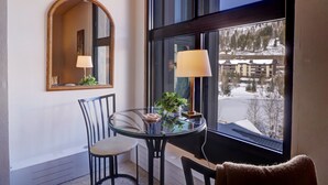 A room with a round glass table, two chairs, a lamp, a mirror, and a view of a snowy landscape through large windows. - Perfectly Located Hotel Room, Steps to American Eagle chair - Ski-In Ski-Out Property - MP518H by RedAwning (Copper Mountain)