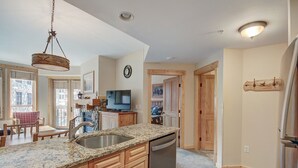 A kitchen with granite countertops, wooden cabinets, a stainless steel sink, and a dishwasher. There is a television on a wooden stand, a clock on the wall, and a wooden door leading to another room. - Taylors Crossing In The Heart Of Copper Center Village - Short Walk to American Eagle/American Flyer - TX312 by RedAwning (Copper Mountain)